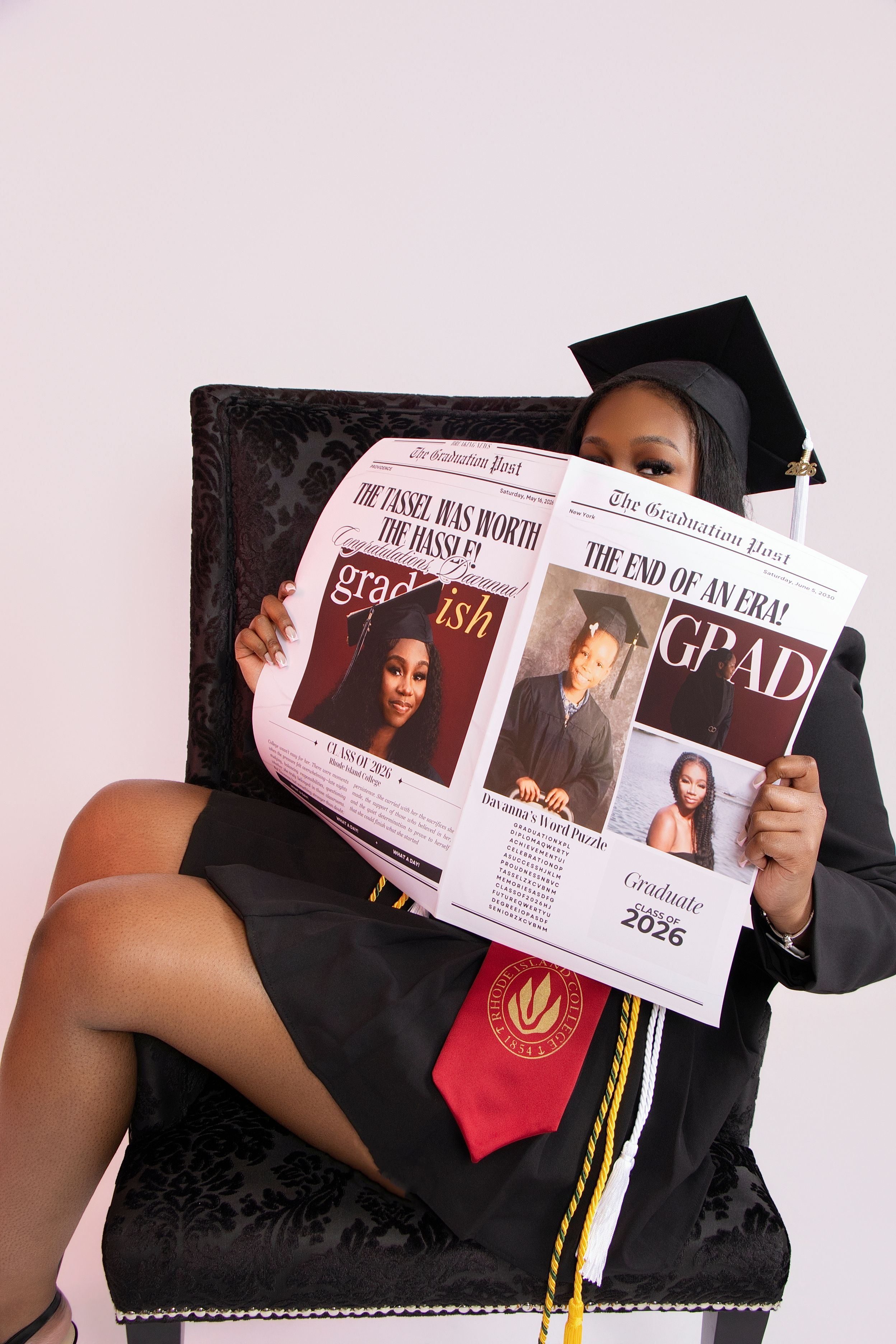 A graduate in cap and gown holds a personalized graduation newspaper while seated in a chair, class of 2026.