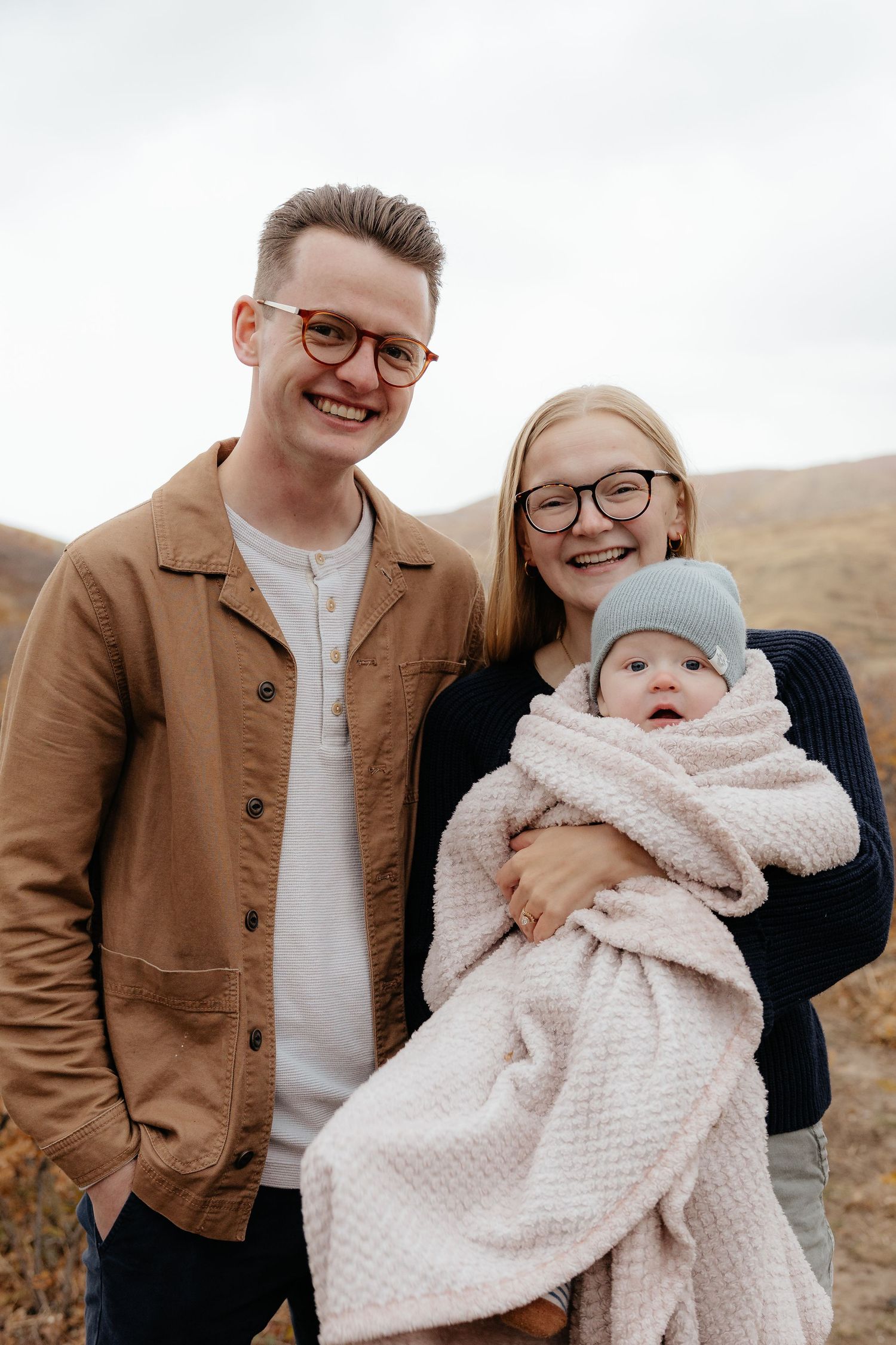 Smiling couple wearing glasses holding their baby wrapped in a pink blanket in an autumn landscape.