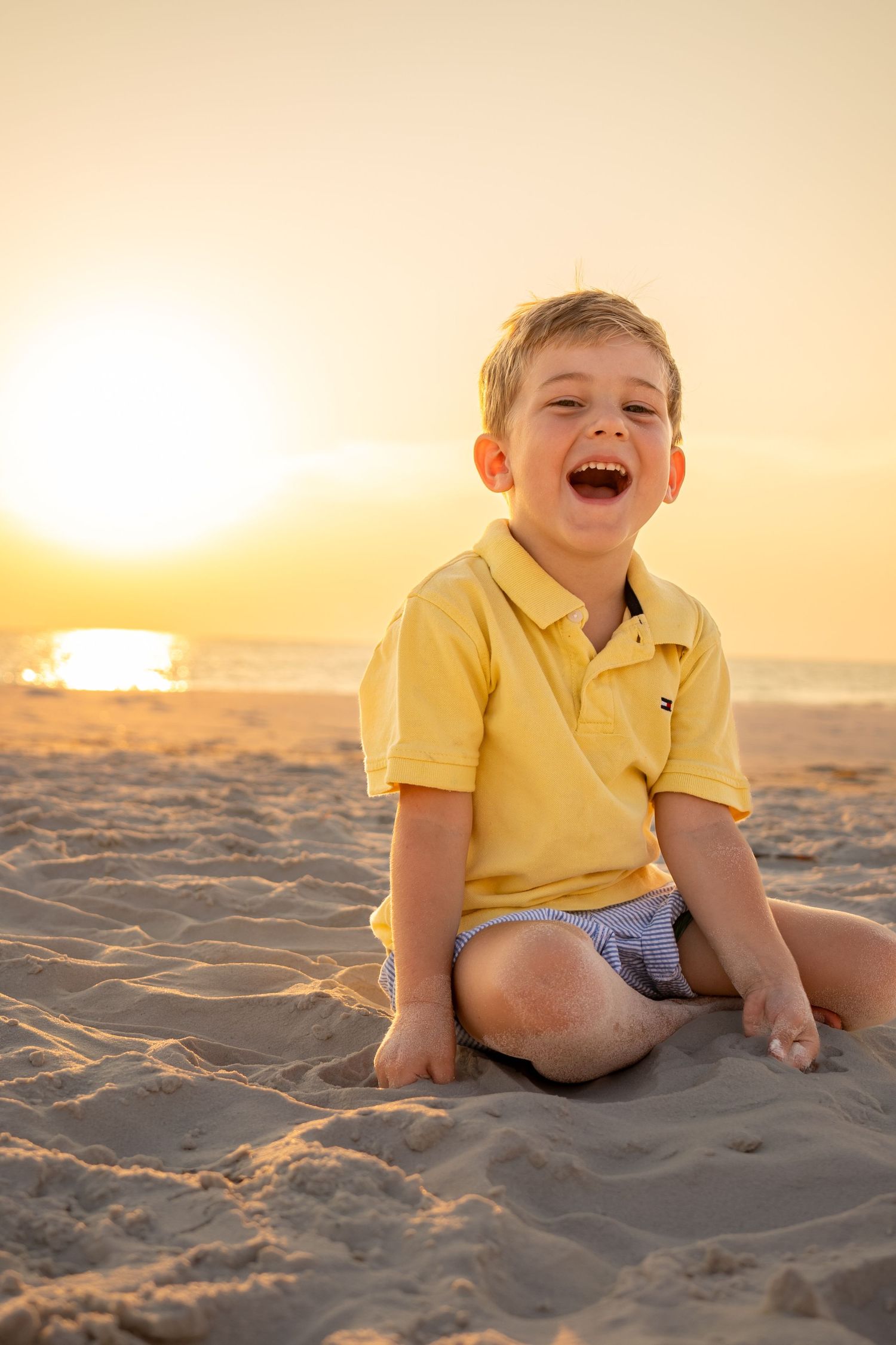 A child in a yellow polo shirt laughs joyfully while sitting on a sandy beach at sunset.