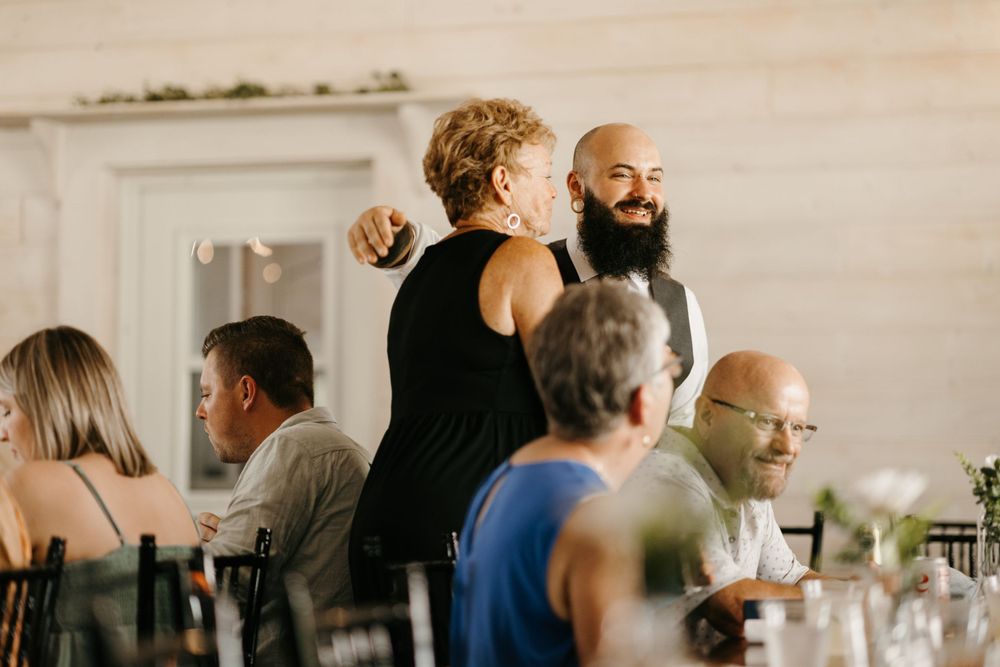 Bearded groom in vest laughs with wedding guests during reception dinner.