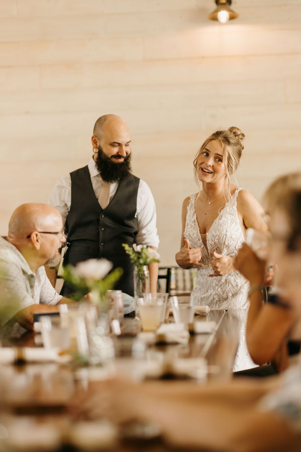 Bride in lace gown and bearded groom smile at wedding reception dinner table.