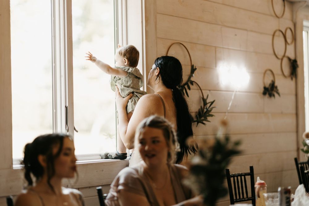 Woman holds toddler near window at rustic wedding reception venue.