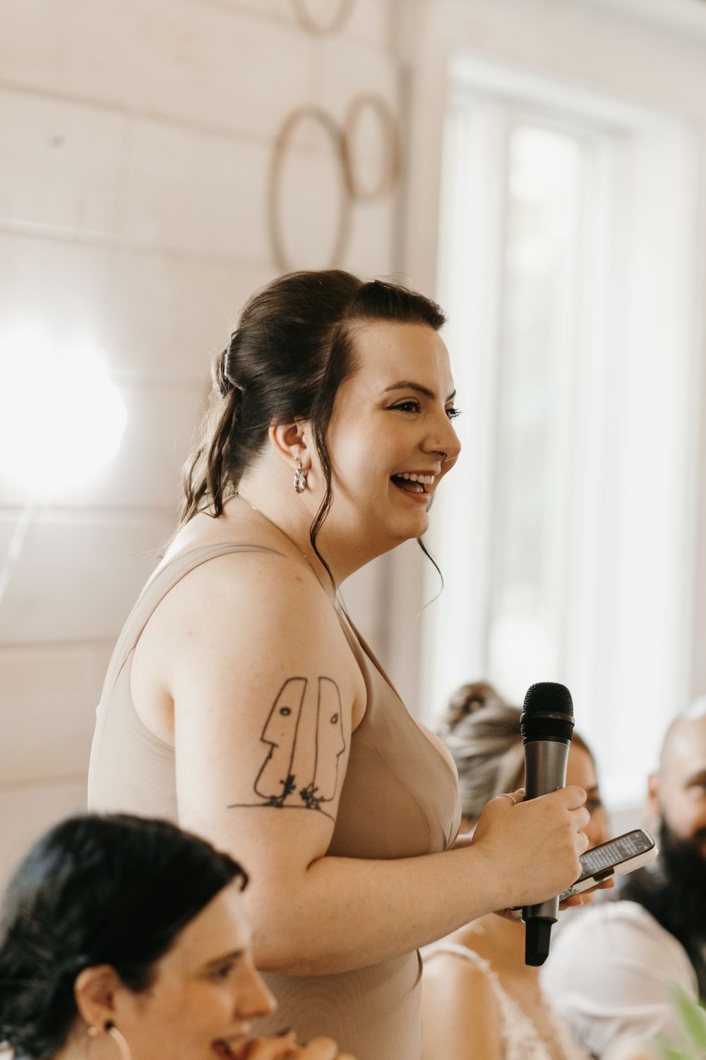 Bridesmaid with tattoo gives speech holding microphone at wedding reception.