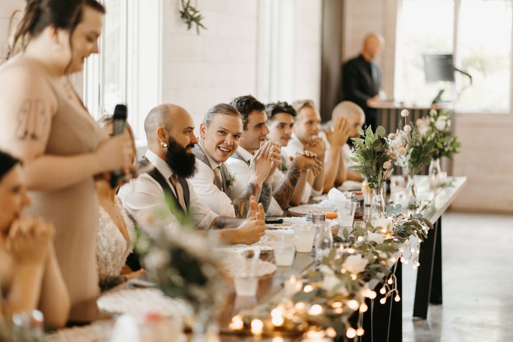 Wedding party guests applaud and smile at decorated reception head table.