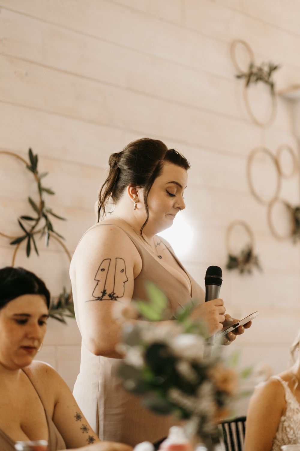 Tattooed bridesmaid reads speech from phone while holding microphone at reception.