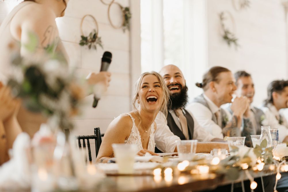 Bride and bearded groom laugh joyfully during wedding reception speech.
