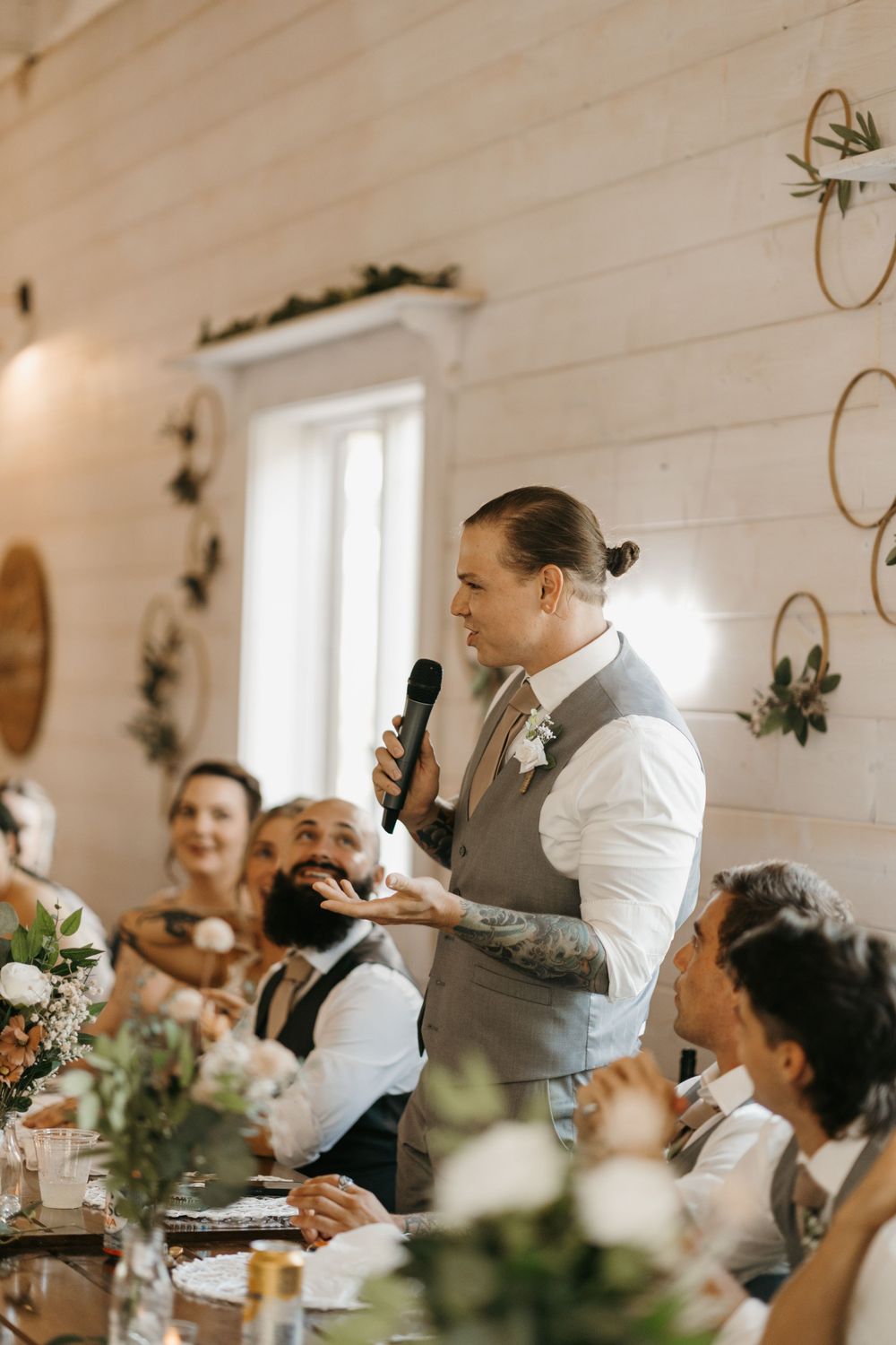 Tattooed groomsman delivers heartfelt wedding speech with microphone at elegant barn reception.