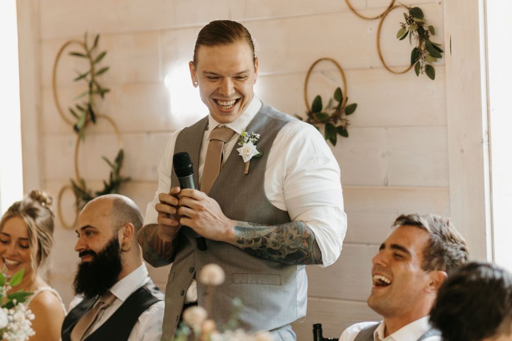 Smiling tattooed best man gives wedding speech while guests laugh at reception.