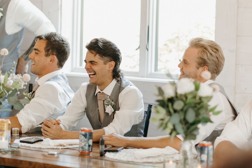 Three men laugh joyfully at wedding reception table with floral centerpieces.