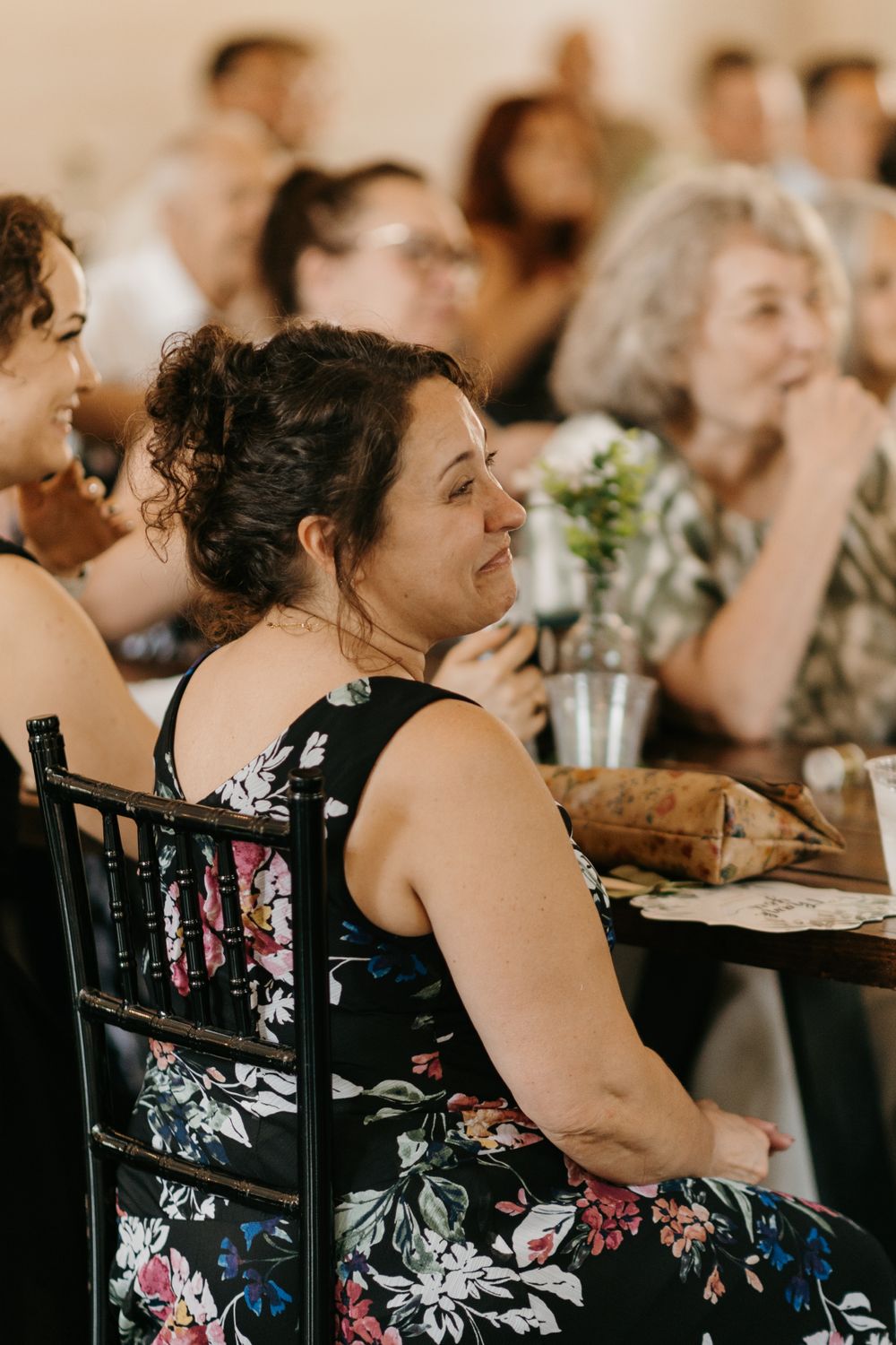 Woman in floral dress smiles warmly while seated at wedding reception.