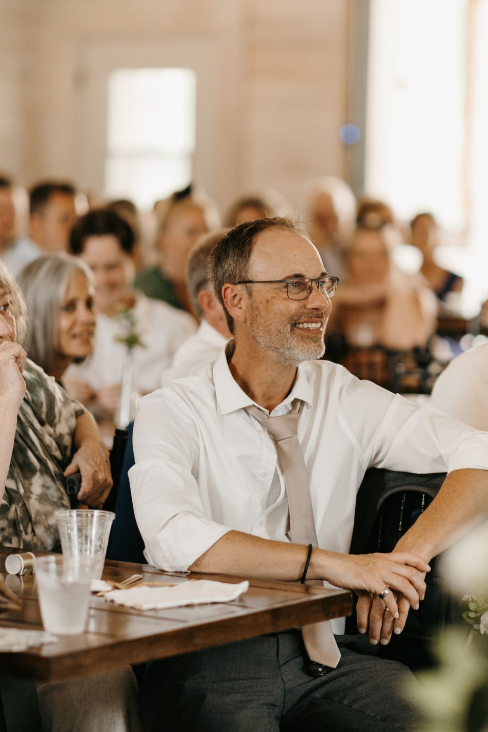 Smiling man with glasses relaxes at wooden table during wedding reception.