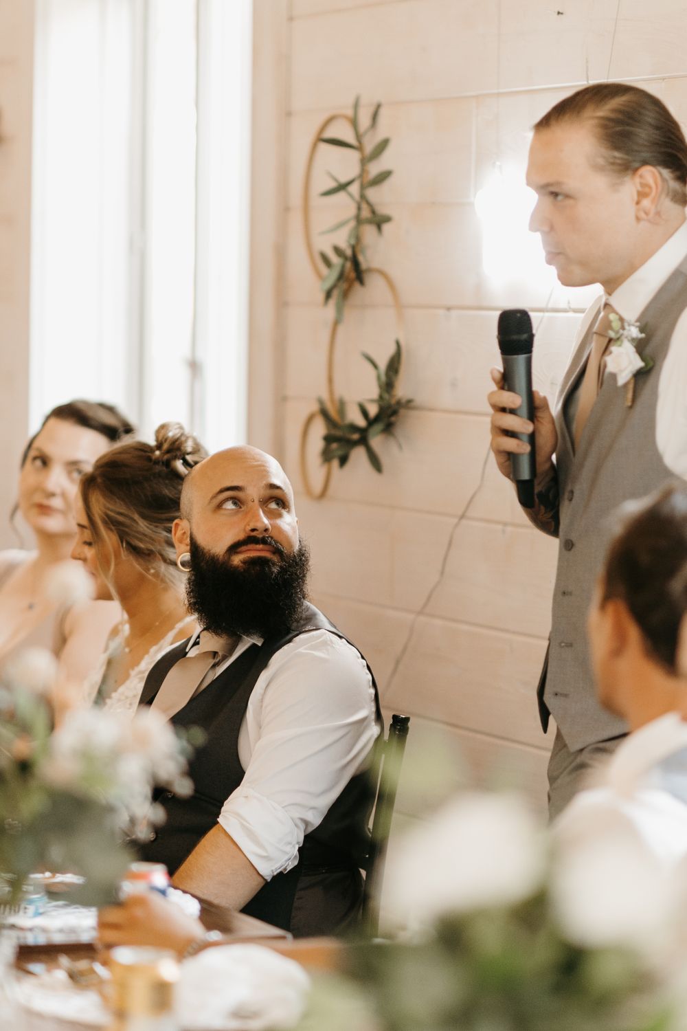 Groomsman delivers wedding speech while bearded groom listens at head table.