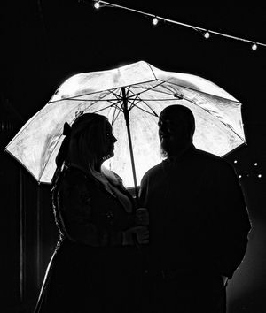 Romantic black and white silhouette of bride and groom under umbrella with string lights.
