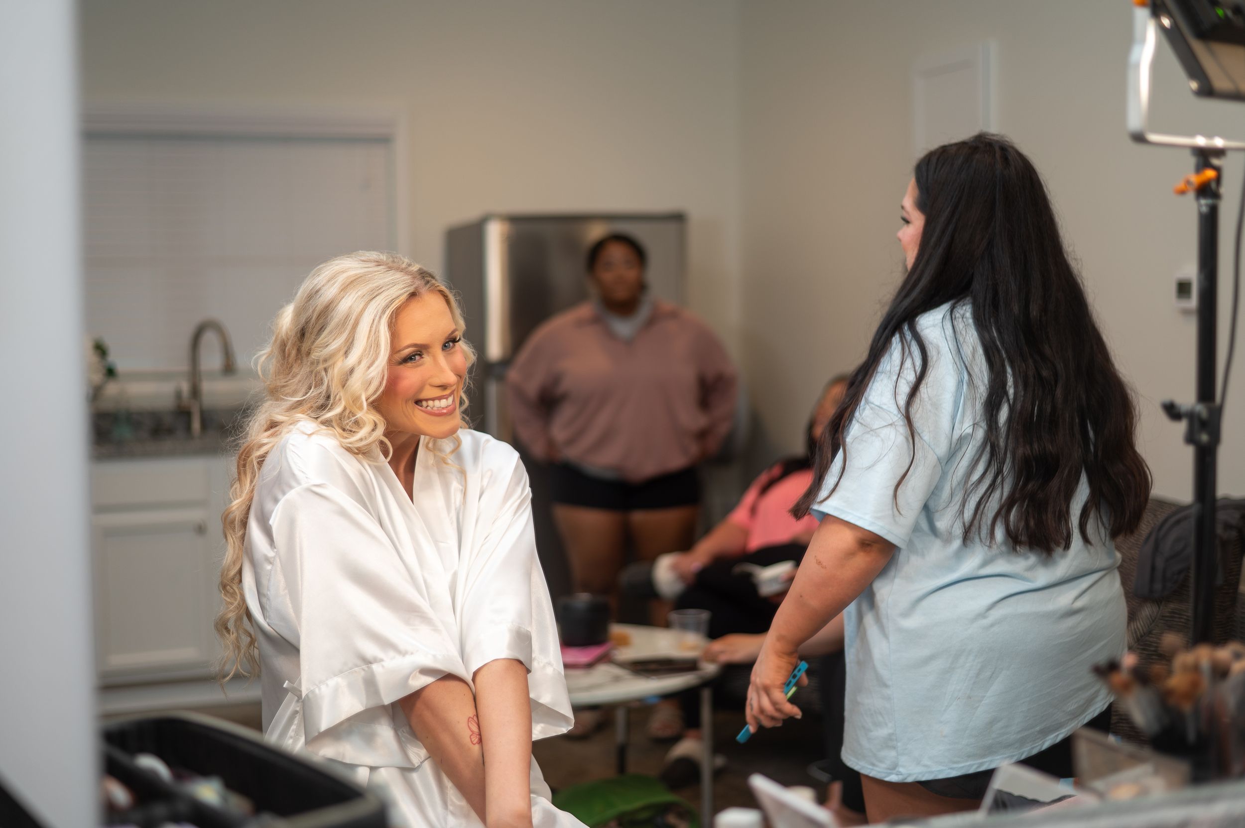 Bride smiling in white robe during wedding preparation with makeup artist and guests in background.