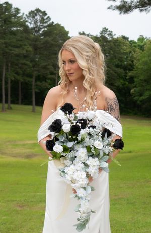 Blonde bride in white off-shoulder gown holding black and white floral bouquet outdoors.