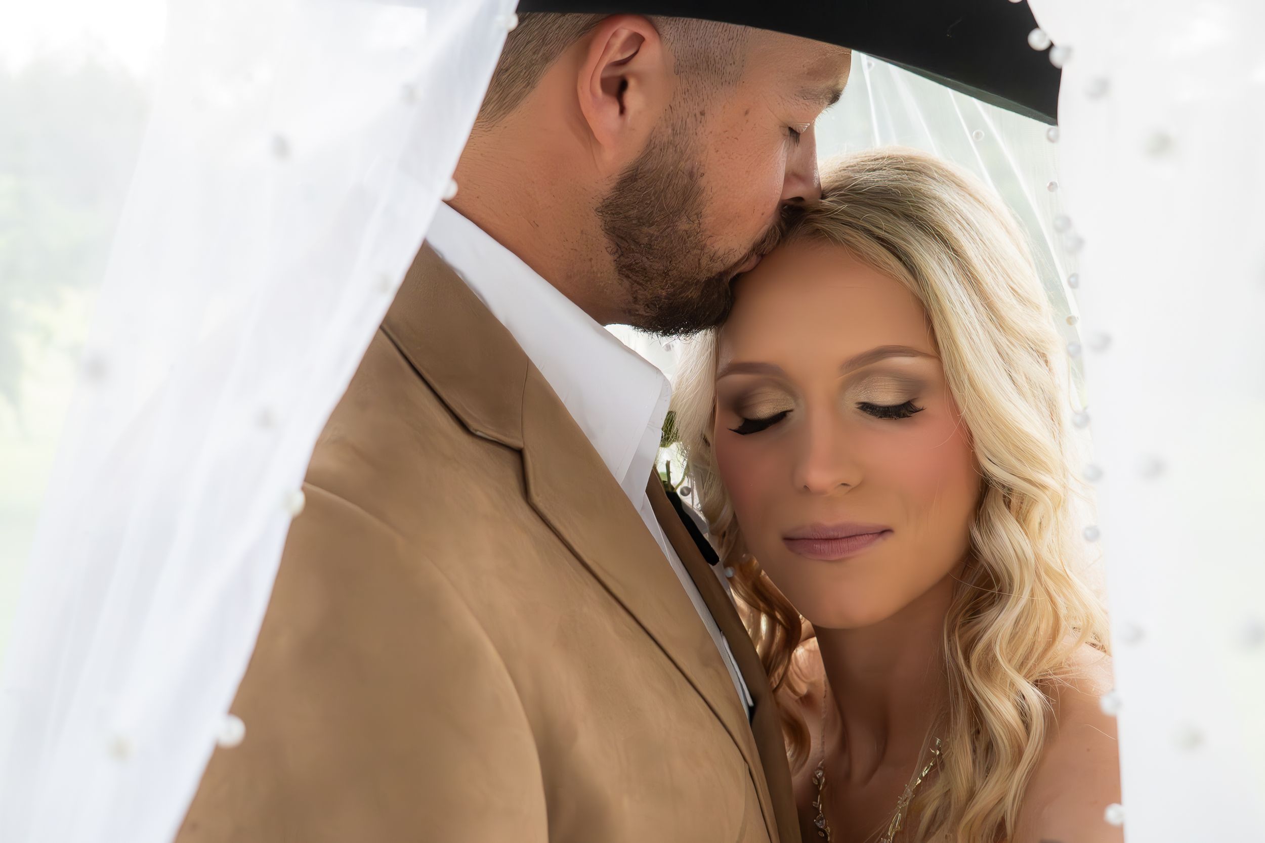 Groom tenderly kissing bride on forehead beneath a sheer veil in soft natural light.