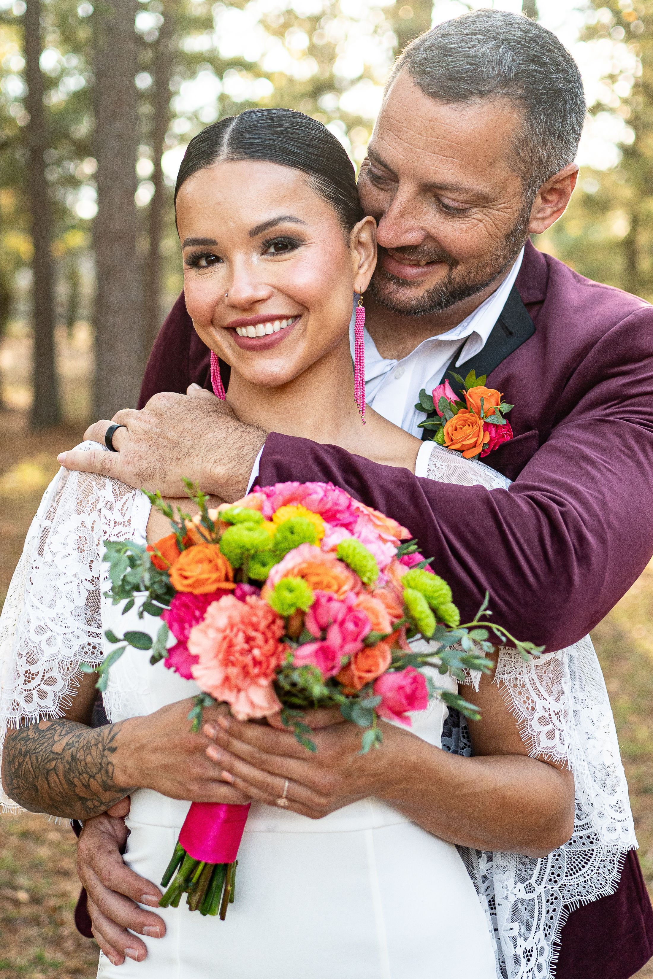 Happy bride and groom embracing in pine forest with colorful orange and pink bouquet.