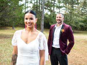 Groom in burgundy velvet suit smiling at bride in white lace dress during first look.