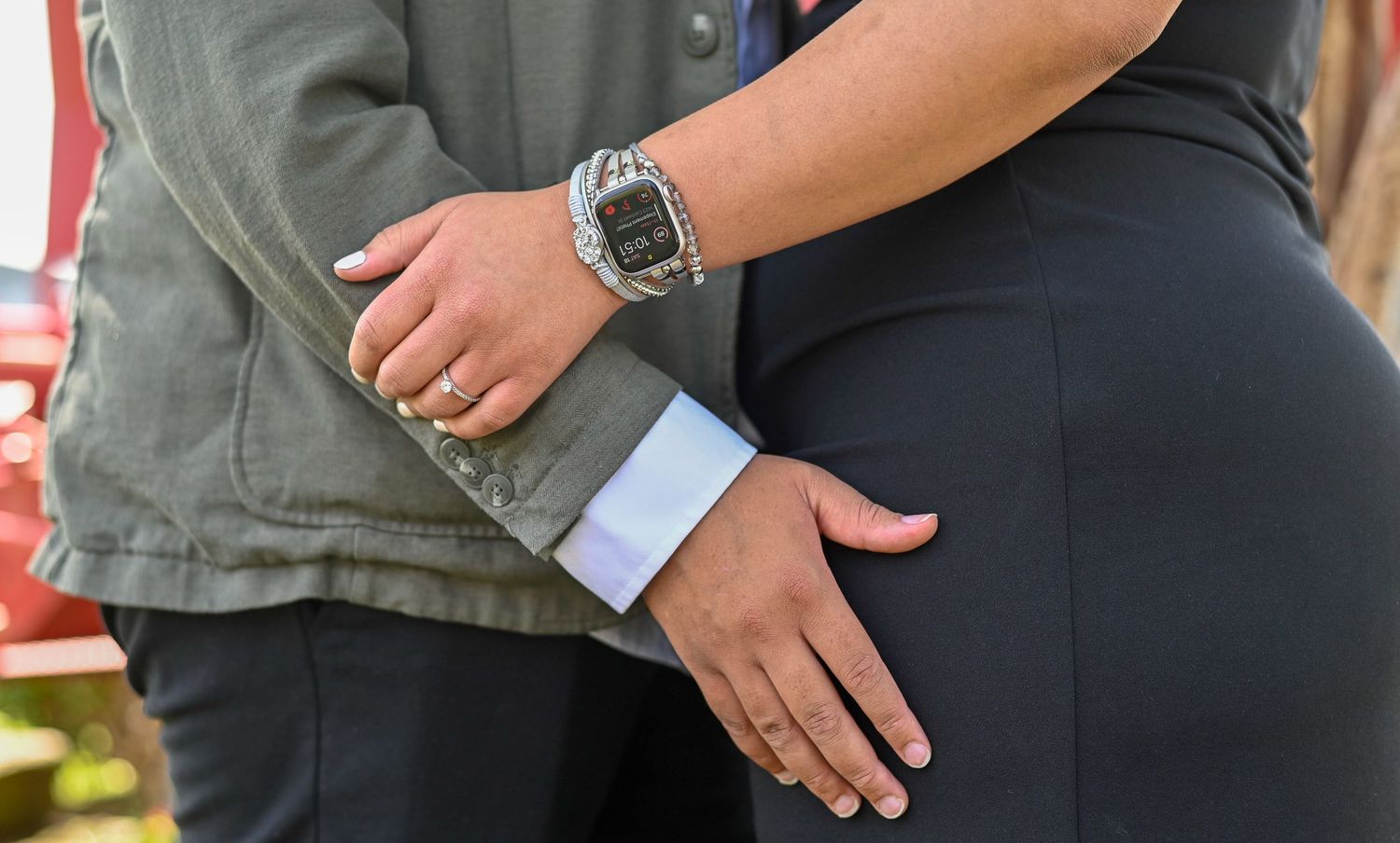Close-up of a couple holding hands, woman wearing a smartwatch and engagement ring, man in a gray jacket.