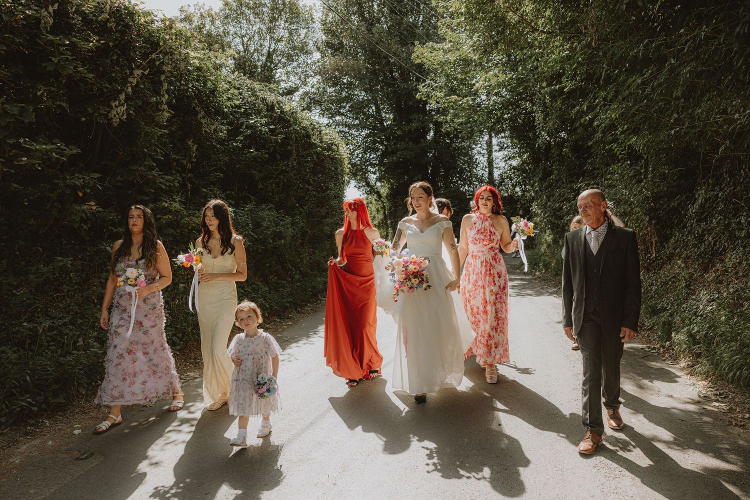 Wedding guests walk along a tree-lined path in formal attire during an outdoor celebration.