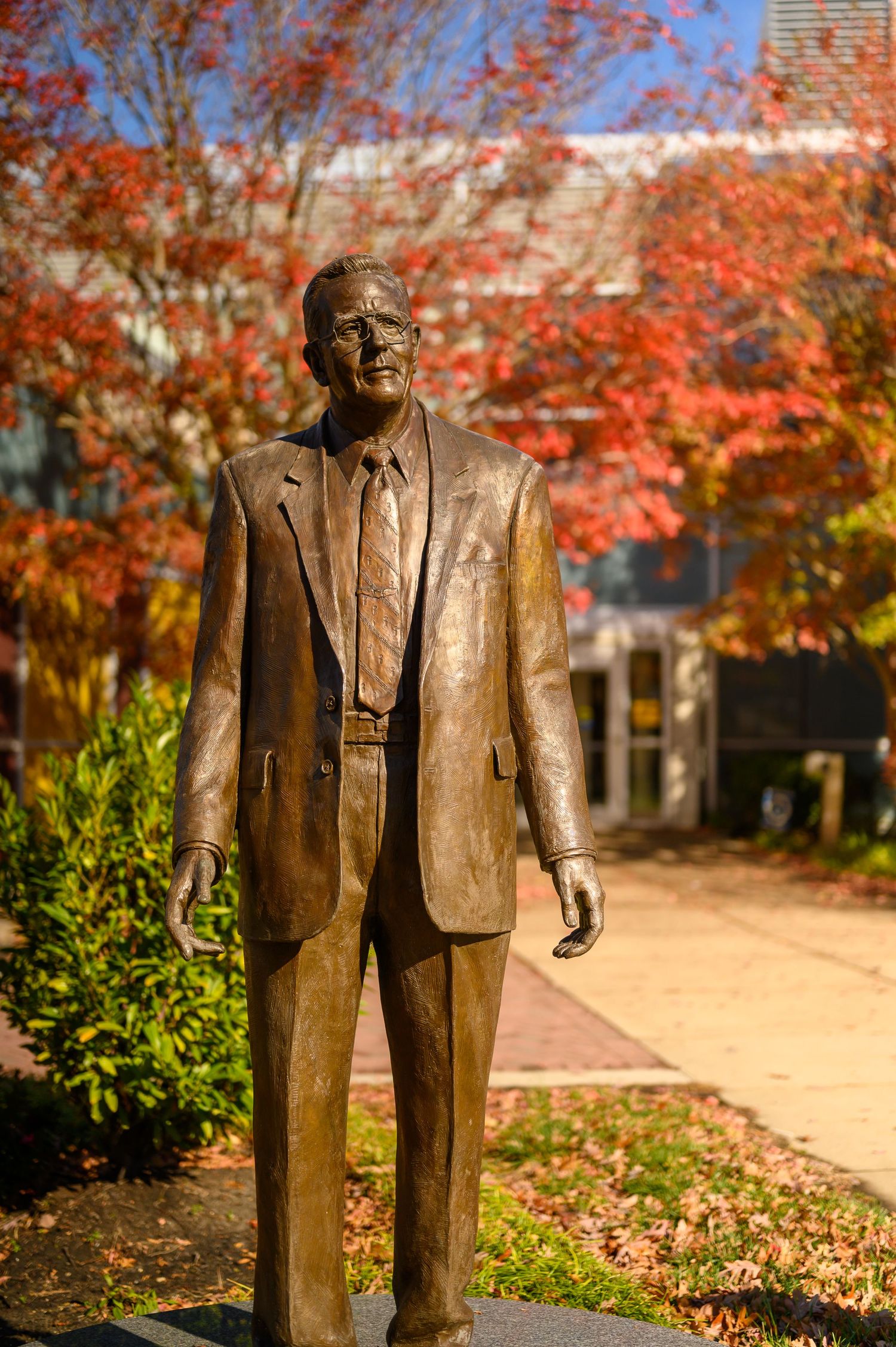 Bronze statue of a man in a suit stands against vibrant autumn foliage.