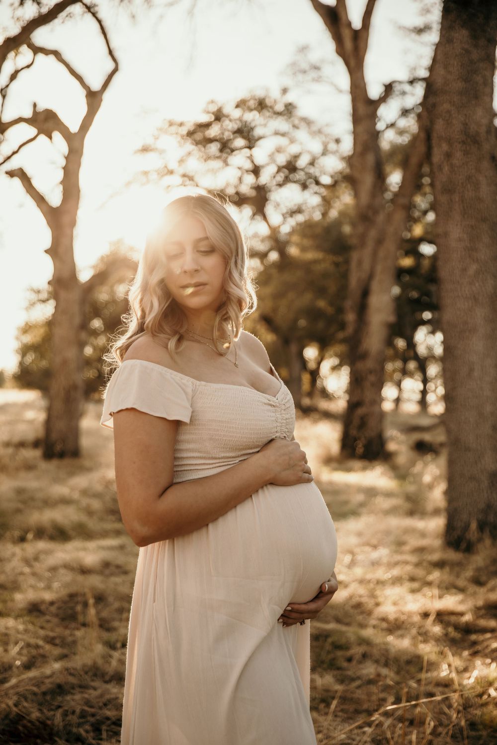 Soft sunlit maternity portrait in cream dress among oak trees at golden hour.