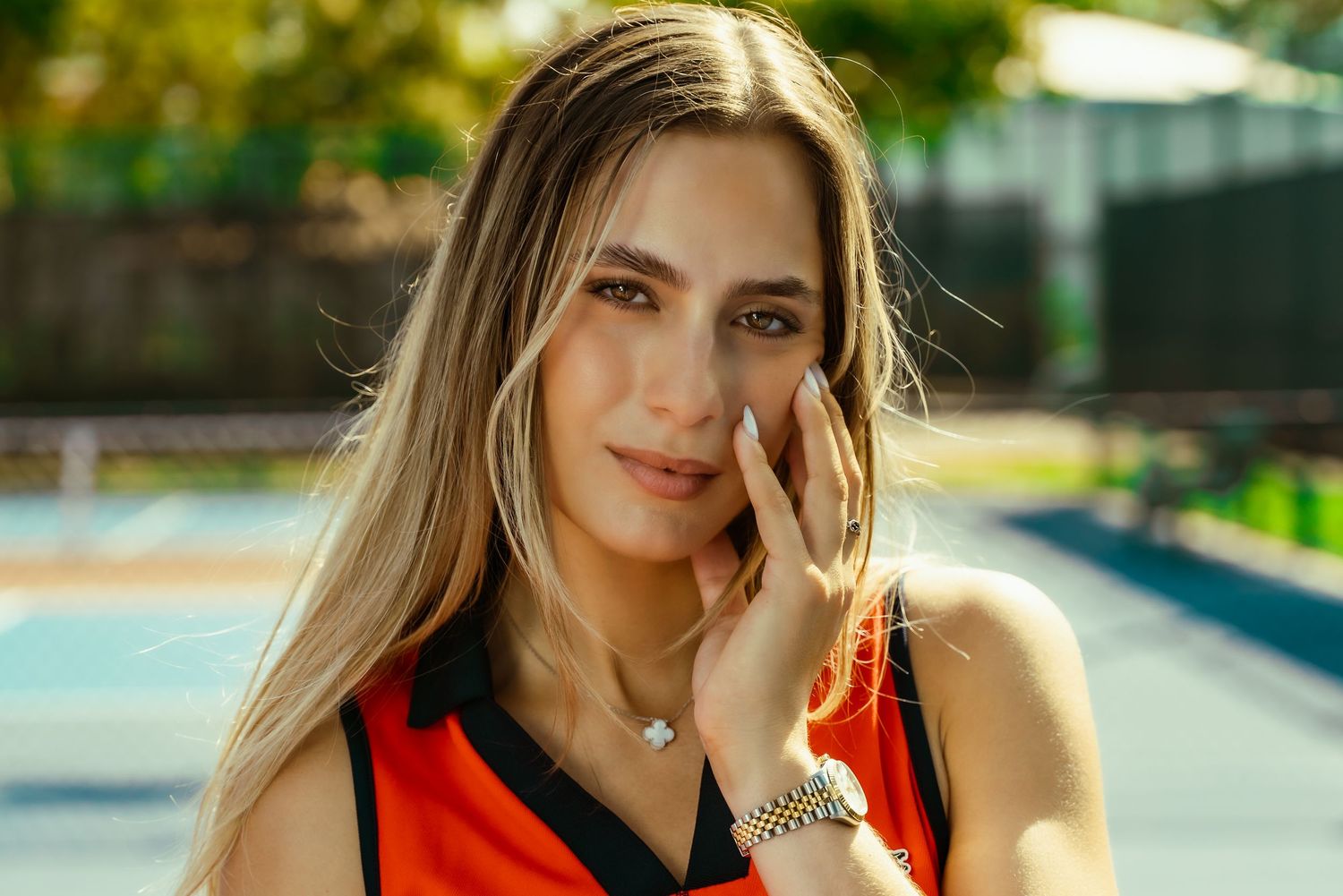 Tennis player in orange athletic top poses outdoors on sunny court.