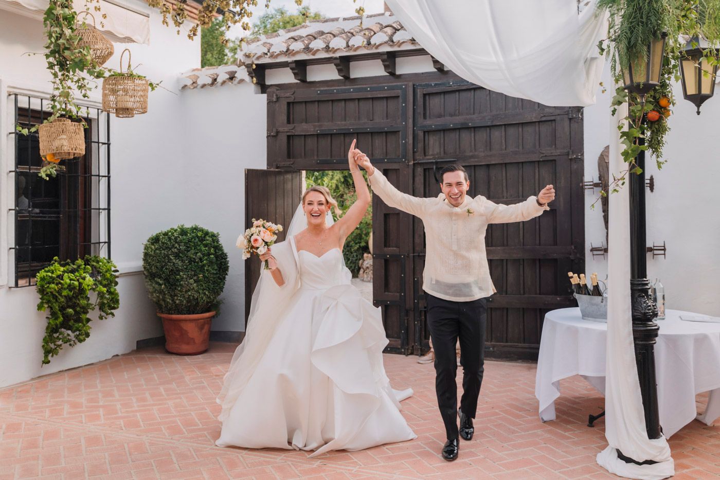 A joyful wedding celebration at a Spanish-style courtyard with white decorations and potted greenery.
