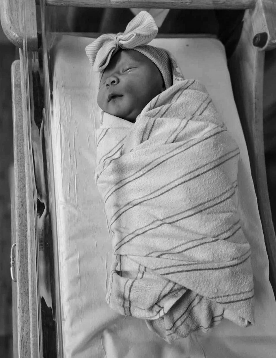 A black and white photograph shows a newborn baby swaddled in white cloth with a bow on their head lying down.