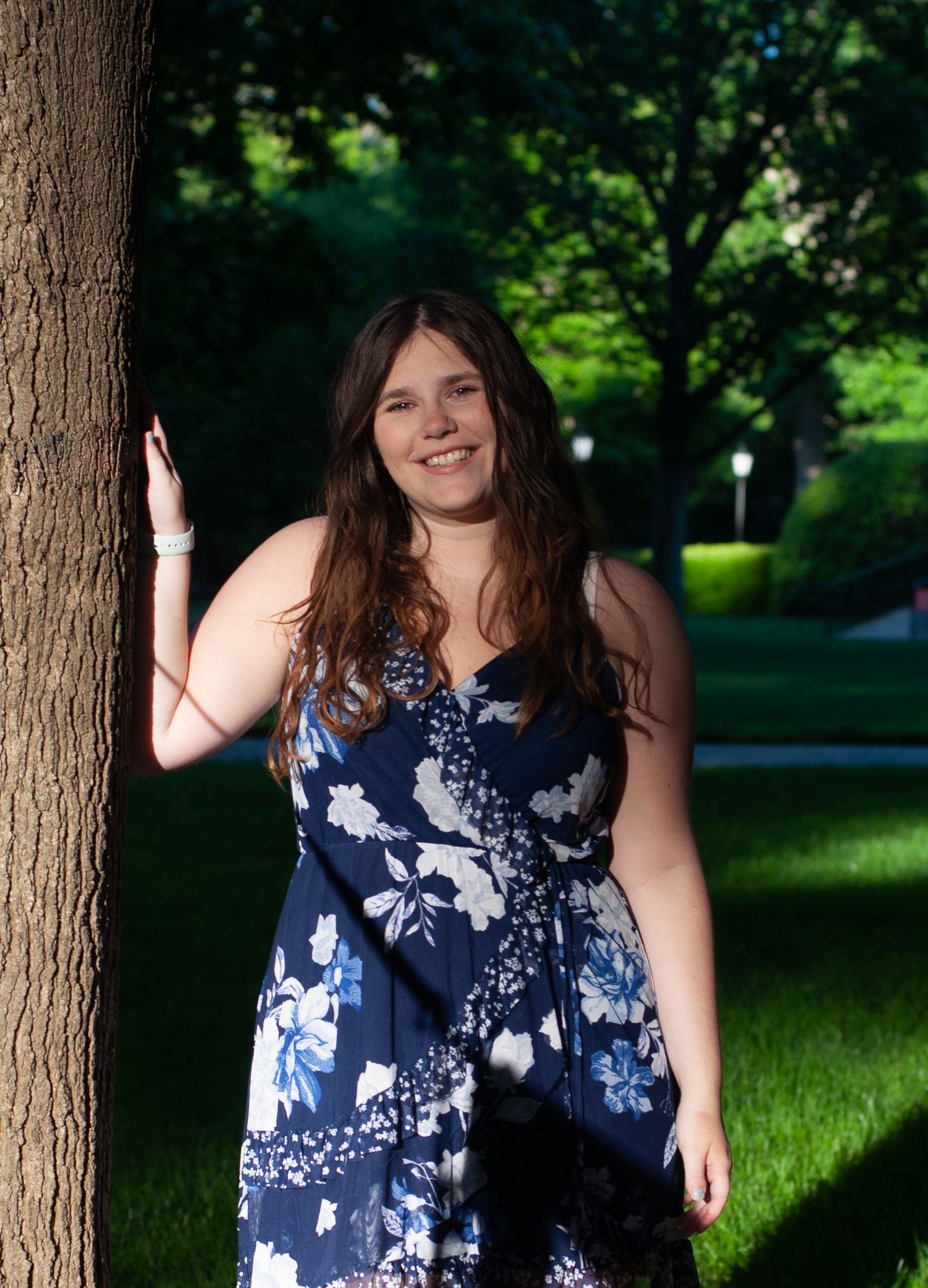 College graduation photo of a young woman standing beside a tree on her campus lawn