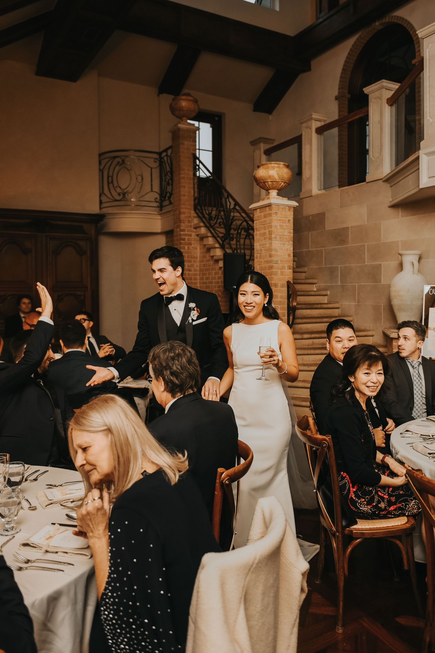 Wedding guests seated at elegant round tables in a rustic venue with stone walls and wooden beams.