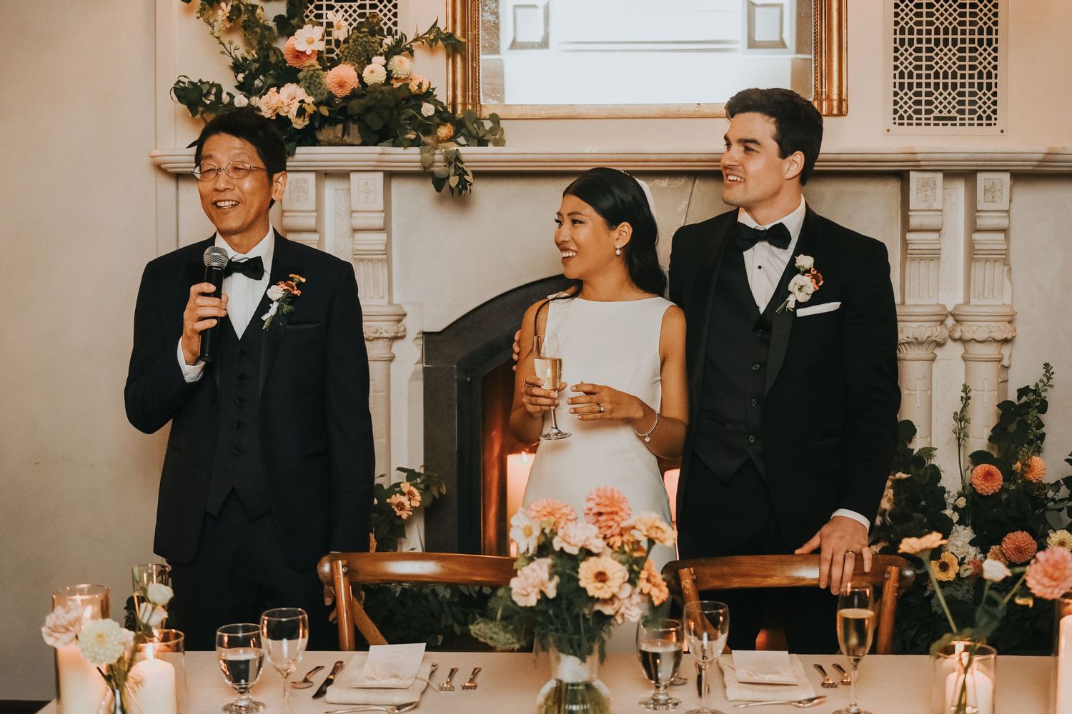Wedding party members in formal attire stand at an elegantly decorated reception venue during speeches.