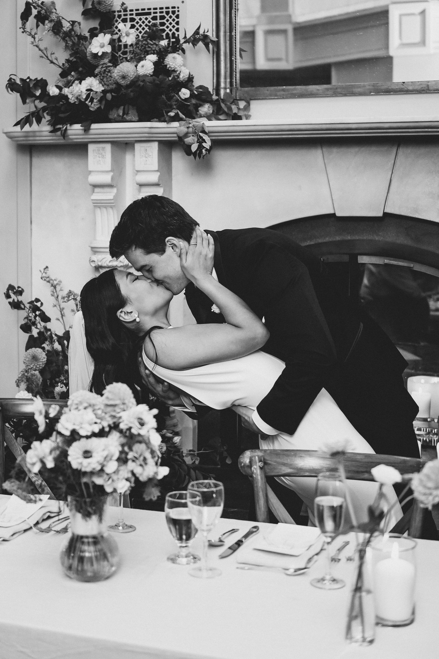 A romantic couple shares a passionate kiss at an outdoor dinner table decorated with wine glasses and floral arrangements.