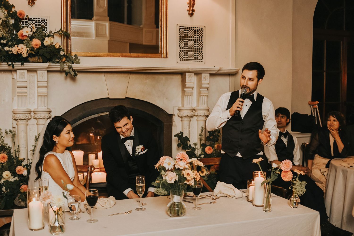 Candlelit wedding reception features a speaker at head table with elegant floral arrangements and white decor.