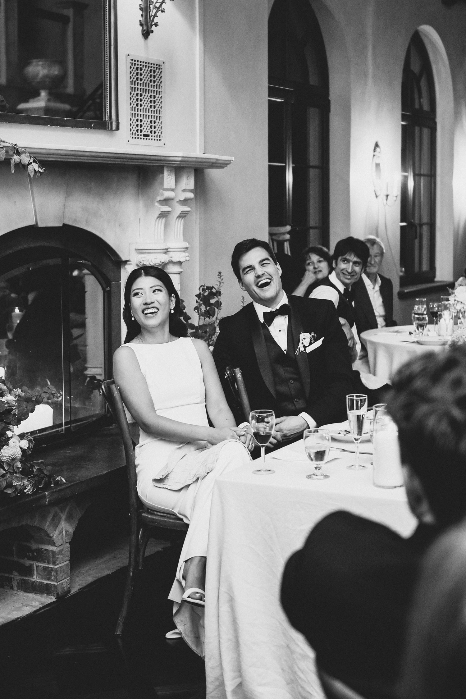 Candid black and white photograph capturing joyful laughter during a wedding reception at an elegant venue with arched doorways.