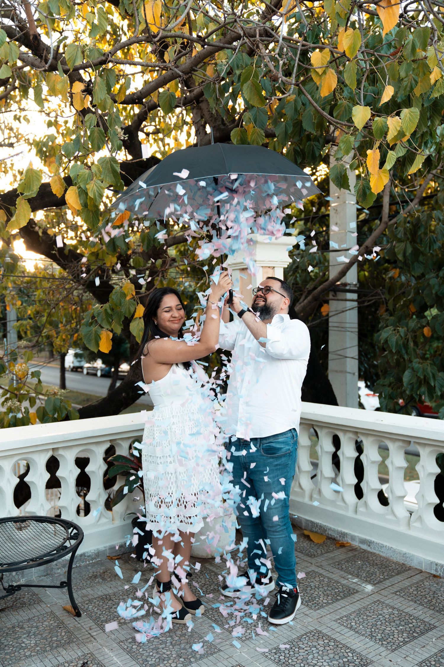 Joyful couple celebrates under umbrella with confetti on elegant white balcony.