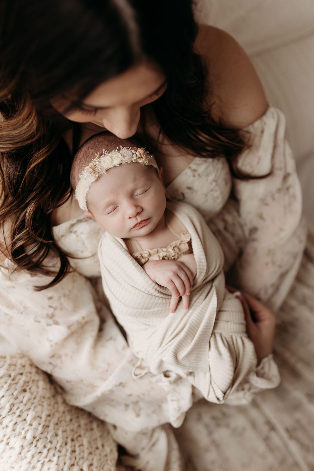 Mother in floral dress tenderly kisses sleeping newborn wrapped in cream swaddle with delicate floral headband.