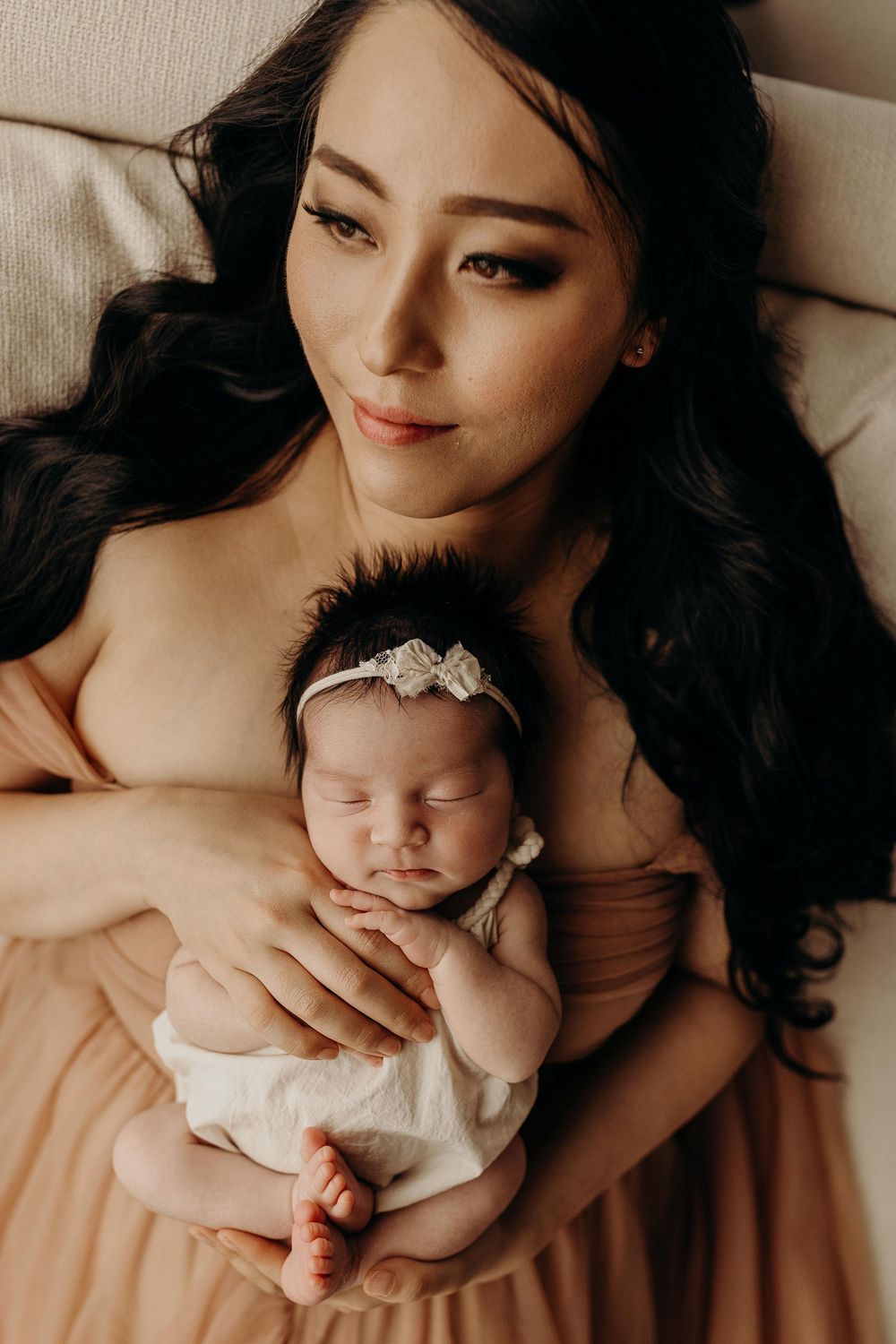 A mother with long dark hair in a peach dress tenderly holds her sleeping newborn baby with a floral headband.