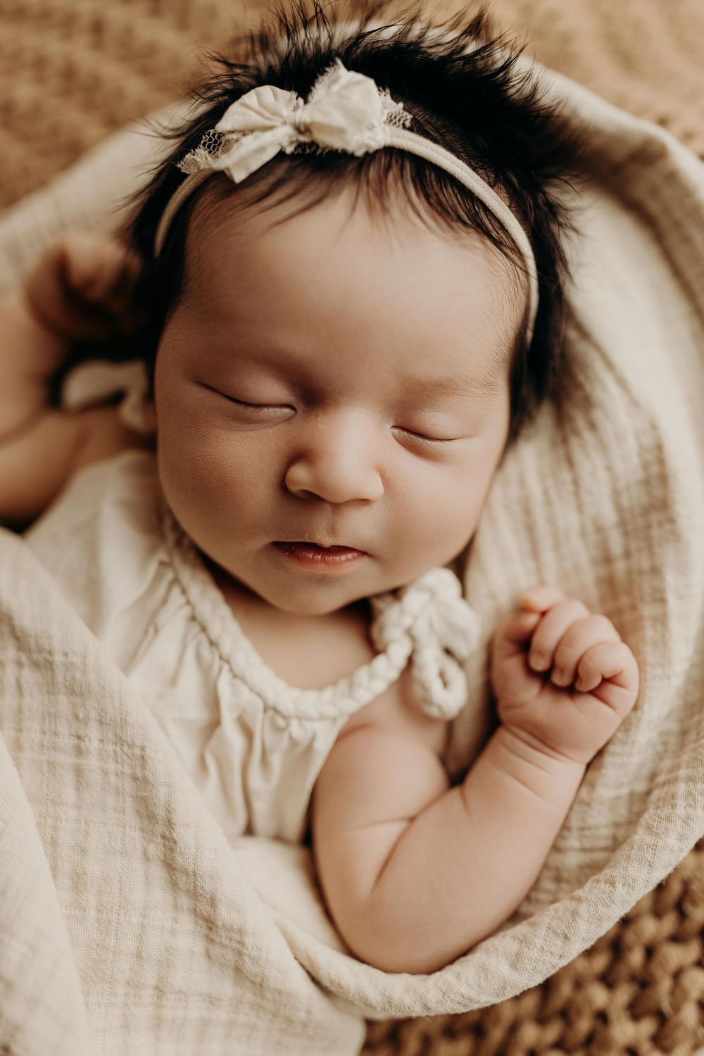 Sleeping newborn baby girl wearing a white bow headband, wrapped in a cream muslin blanket.