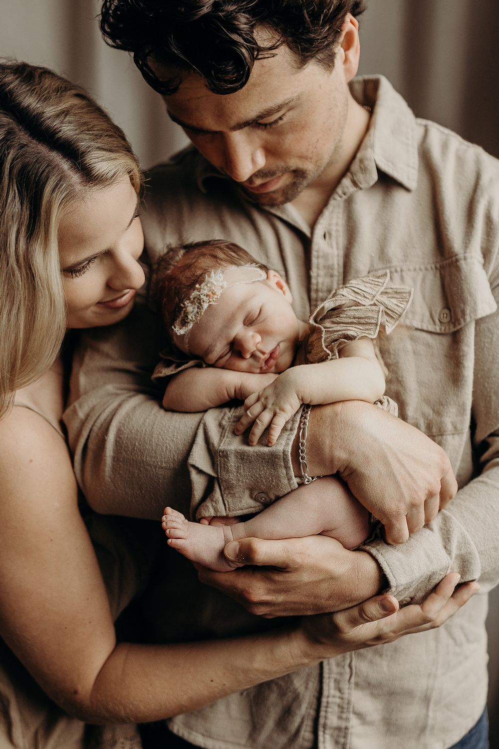 New parents tenderly cradling their sleeping newborn baby dressed in beige, sharing a loving family moment.