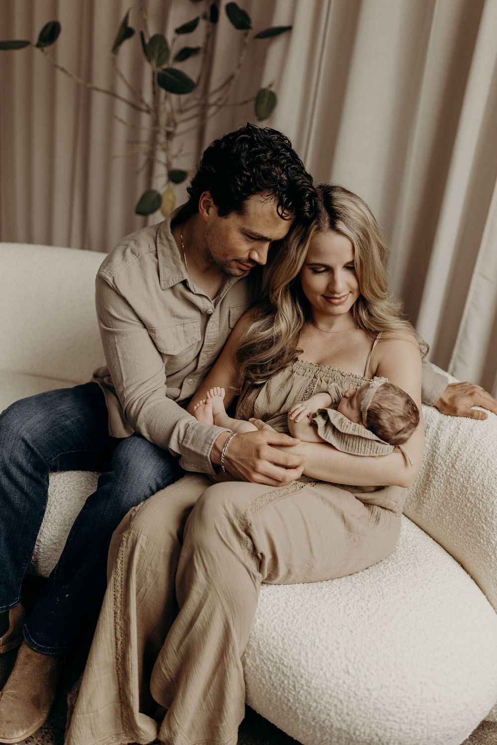 A couple sits on a white sofa, lovingly gazing at their newborn baby in a warm, neutral-toned nursery setting.