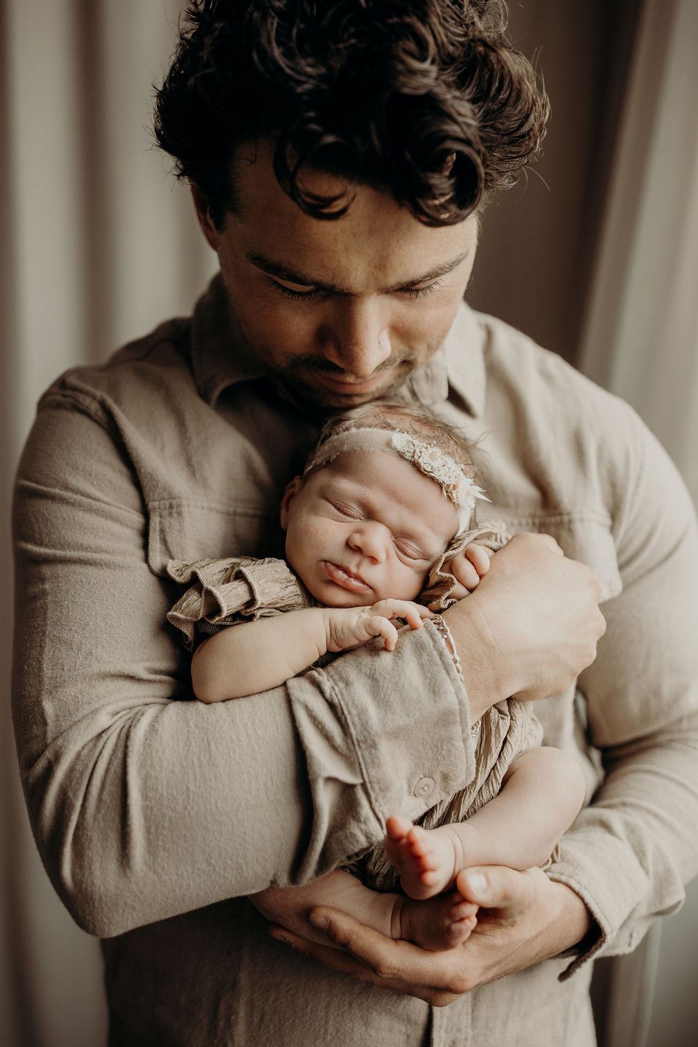 A tender father cradles his sleeping newborn baby, both dressed in neutral tones, sharing a peaceful bonding moment.