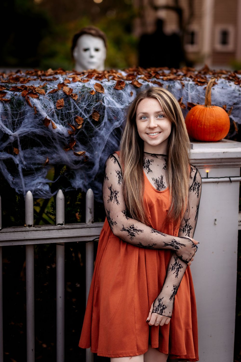 Halloween scene with orange dress, spooky lace sleeves, cobwebs, pumpkin and white mask decoration in background.