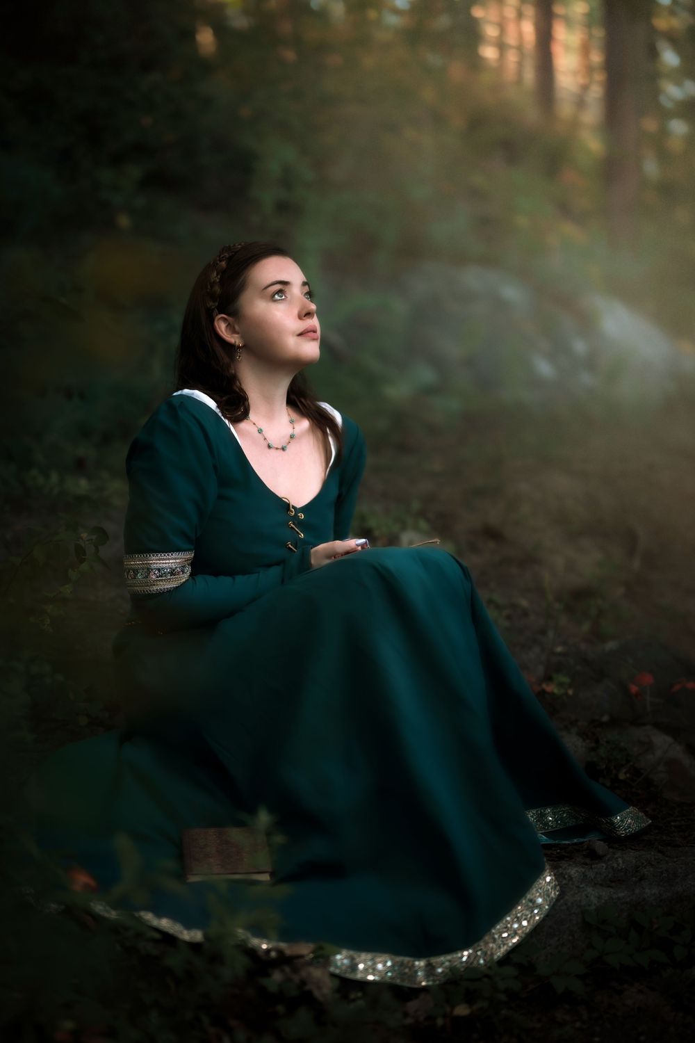 A person in a teal dress sits gracefully on forest ground during a moody autumn photoshoot.