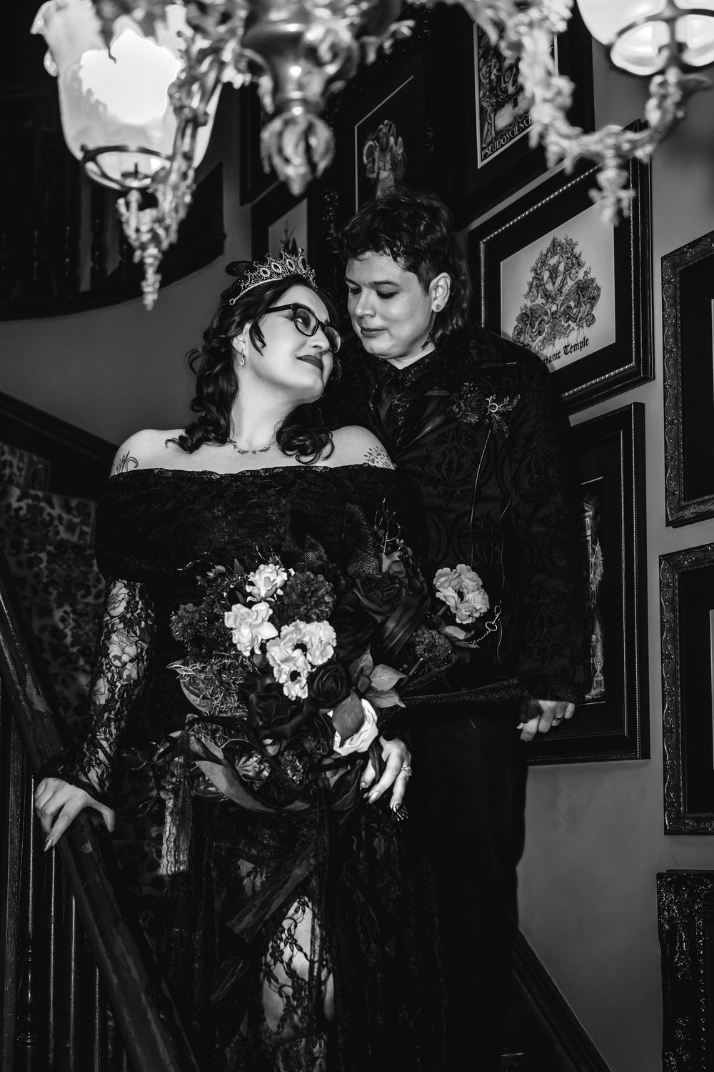 Gothic wedding couple in black attire on staircase, bride in lace gown with tiara holding bouquet, black and white photo.
