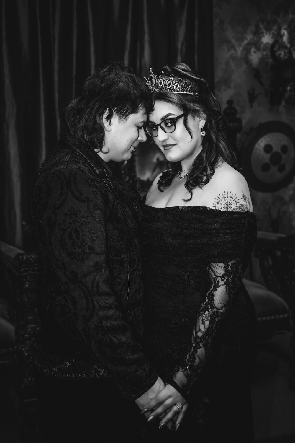Black and white photo of a couple in elegant dark attire, woman wearing a crown and glasses, sharing an intimate moment.