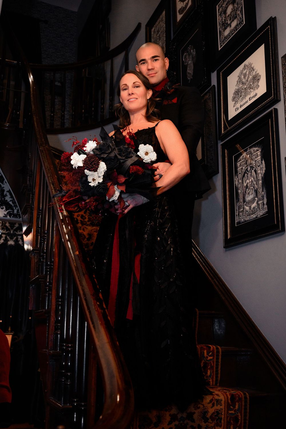 A couple poses on an ornate wooden staircase with black railings surrounded by framed artwork on the walls.