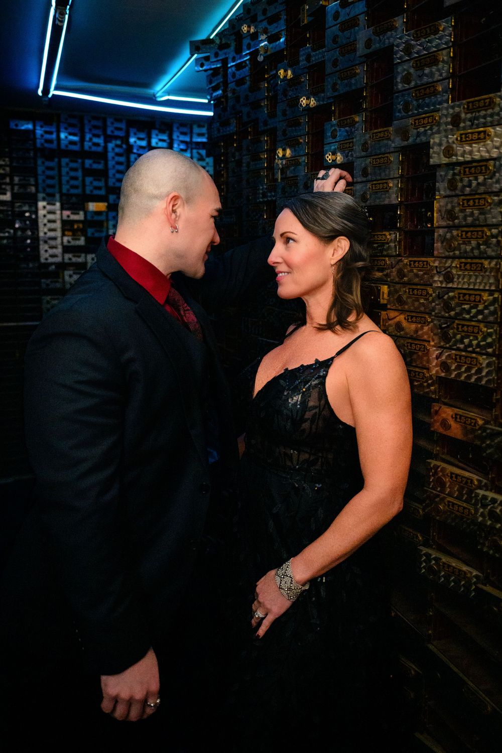 A couple in formal attire share an intimate moment against a dark brick wall with blue neon lighting.