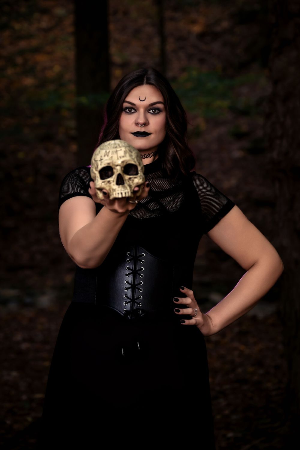 A gothic-styled figure in black clothing poses dramatically with a human skull in a dark forest setting.