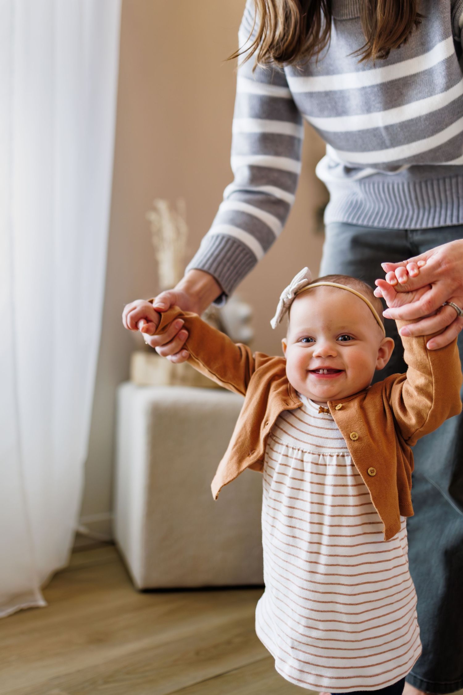 A baby in a striped dress and tan cardigan taking first steps while being supported.